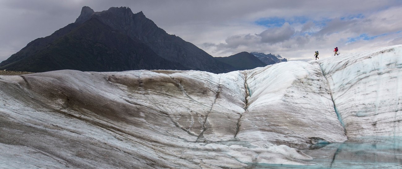 Two hikers traversing a glacier with mountains in the background
